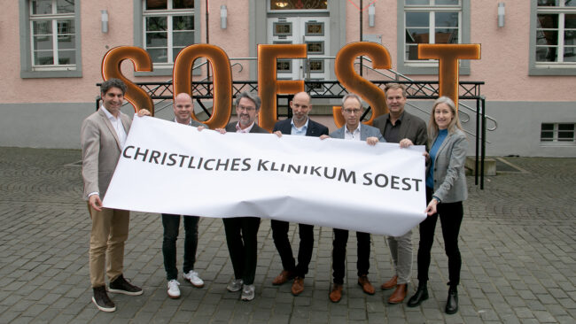 Fortschritte Krankenhaus-Fusion Das Bild zeigt (v.l.): Christan Schug (Geschäftsführer KlinikumStadtSoest), Tobias Franke (Kfm. Direktor Marienkrankenhaus Soest), Dr. Eckhard Ruthemeyer (Bürgermeister Stadt Soest), Christian Larisch (Geschäftsführer Hospitalverbund), Guido Ponto (Vorsitzender Verwaltungsrat Hospitalverbund), Thorsten Roy (Geschäftsführer Christliches Klinikum Unna) und Sabine Boschert (Kfm. Direktorin KlinikumStadtSoest). Sie halten gemeinsam ein Banner mit der Aufschrift "Christliches Klinikum Soest".